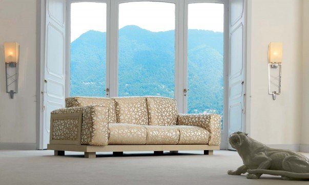 This picture shows a luxurious living room with a white leather sofa set in the center. The walls are a light yellow, with a patterned wallpaper lining the upper half of the wall. The floor is a mix of dark and light hardwood planks. On the left side of the room is a modern fireplace with dark marble accents, flanked by two tall glass windows. On the right is a built-in shelving unit, with a flat-screen television mounted above it. A round coffee table is placed in the center of the room, topped with a decorative vase filled with white