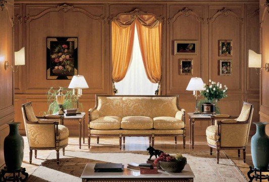 This picture shows an open, modern living room with an exposed brick wall. The floor is covered with a light-colored carpet and there is a white sofa and two armchairs in the center of the space. The room also has two gray accent chairs and a large window with closed shutters. The walls and ceiling are painted white, and there are two decorative lamps on either side of the sofa. At the back of the room is a sliding glass door that leads to an outdoor patio area.