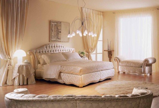 The picture shows a luxurious bedroom with a distinct modern design. The walls are covered in a blue and white diamond patterned wallpaper, and there is a large white four-poster bed in the center of the room. Above the bed hangs a matching white chandelier, and there is a navy blue colour scheme in the furniture pieces and decorative accents throughout the room. A small bookshelf at the far end of the room is filled with books and other objects while two white nightstands frame the bed.