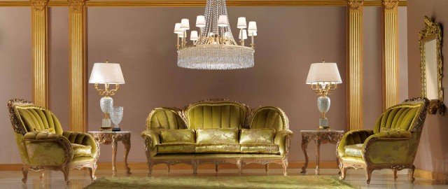 This picture shows an ornate and luxurious dining room with a crystal chandelier descending from a gold-plated ceiling. The walls and floors are decorated with intricate details, including detailed wall artwork and patterned tiling. A large white marble table sits on a large plush rug in the center of the space, surrounded by comfortable and stylish chairs. The room is lit by multiple wall sconces and candelabras, and two large windows offer plenty of natural light.