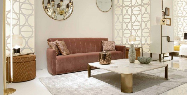 This picture shows a modern and luxurious living room interior. It features white walls and ceiling, as well as a grey floor with a light-colored rug. There is an off-white tufted couch with two yellow accent pillows and a round glass-top coffee table in the center. The fixtures are made of brass and gold, including a round chandelier and standing lamps. There is also a large abstract painting on the wall, adding a touch of elegance to the space.