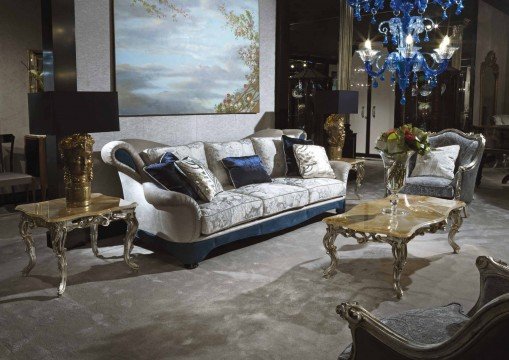 This picture shows a luxurious and modern dining space. There is a white marble dining table surrounded by wooden chairs with ivory seats and tufted backrests. The table is adorned with a golden centerpiece bowl, surrounded by elegantly crafted golden platters. The walls are painted a soft beige color and the floor is composed of light-colored marble tiles. A large chandelier hangs from the ceiling and casts a warm and inviting light over the room.