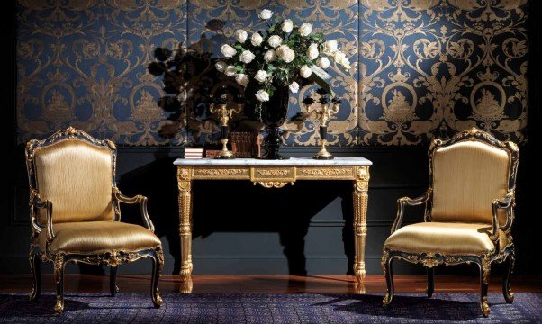 This picture shows a modern luxury living room design. The room is mostly white, with dark hardwood floors and accents of gold throughout. A white leather sectional sofa is centered in the room, with two armchairs upholstered in grey velvet. A gold and white geometric patterned carpet covers the floor in front of the sofa. A large round coffee table with a gold frame is situated in the middle of the seating area. On either side of the seating area are tall glass windows that reach up to the ceiling, providing natural light. On the left side of the room, a