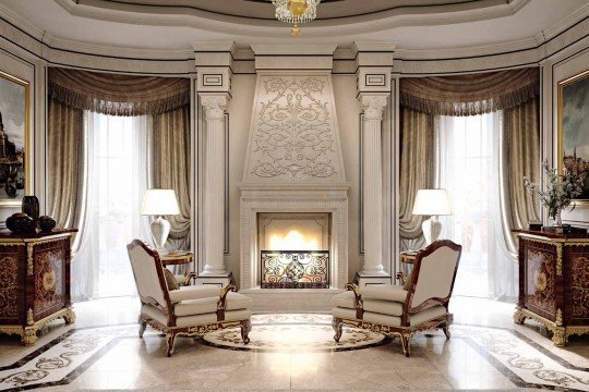 The picture shows a luxurious living room with a classic design. The room features a cream-colored sofa and armchairs covered with red, gold, and teal-colored pillows. A large ornate wooden coffee table rests in the center of the room, and a stately fireplace is visible on the far wall. The walls are painted a pale pink color, and framed artwork hangs on them. Chandeliers are suspended from the ceiling, and several plants are placed throughout the room to add greenery and life.