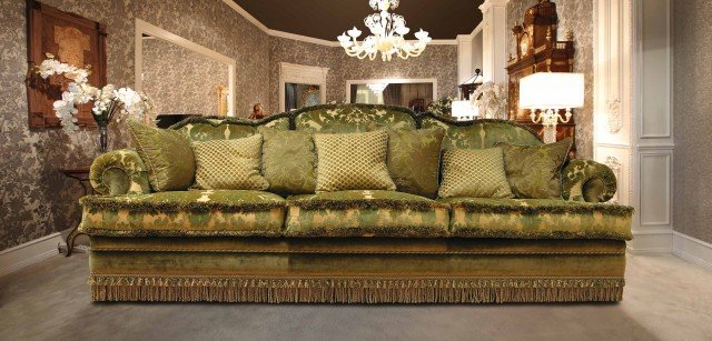 This picture shows a modern, luxurious living room featuring a floral wall mural, a raised hearth fireplace and a black sectional sofa. There is a round wooden coffee table in the center of the room and a patterned rug covering the floor. The walls are painted a light color and the room is accented with dark wood furniture, a sparkling chandelier, and several pieces of decorative art.