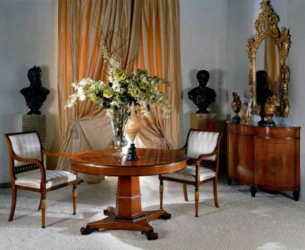 This picture shows a luxurious living room. The room features a dark grey sofa and two armchairs with golden accents, a round ottoman with a black velvet cushion, and a glass-top coffee table. There is an elegant black media cabinet along one side of the wall, and on the opposite side there is a stunning decorative mirror with an intricately carved frame. A white Persian rug covers most of the floor, and additional decorations such as a framed painting and several vases filled with flowers add touches of color.