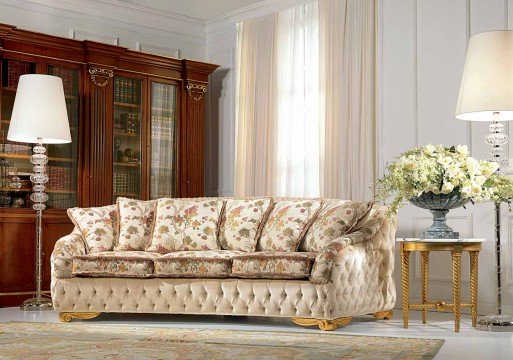 This picture shows a beautifully designed living room with plush, modern furniture and a luxurious gold-plated chandelier. The walls are decorated with white paneling and a bold patterned wallpaper. A light gray couch and armchairs are accompanied by an ornate coffee table and an elegant window treatment. The floor is covered in a white carpet and the room is illuminated by several light fixtures.