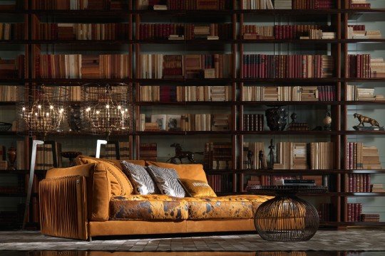 The picture shows a contemporary style living room with several pieces of furniture and decor. There are two white leather sofas, two yellow armchairs, a glass and metal coffee table, two woven ottomans, and a modern wooden sideboard with drawers and shelves. The walls are painted in a light tan color and the floor is covered with a light beige rug. A large abstract painting hangs on the wall above the sideboard, adding a pop of color and texture to the space.