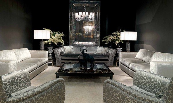 This picture shows a modern, luxury living room. The room is decorated in light and neutral colors, with a white sofa, two neutral-colored armchairs, and an off-white rug. The walls are painted in a dark grey color, and the fireplace features a light grey stone surround. There is a black and white abstract painting on one wall, and a modern metal light fixture hangs from the ceiling.