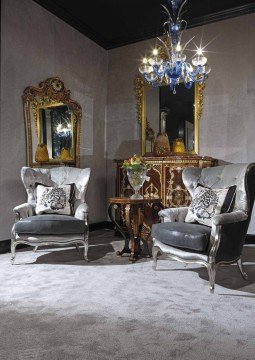 This picture shows a luxurious white and grey living room that is decorated with modern furnishings. The room features a beige couch with a pair of white club chairs, a white coffee table in the center, and a modern fireplace on one wall. The walls are lined with art prints and there is an ornate area rug on the floor. The ceiling is finished with crown molding and recessed lighting.