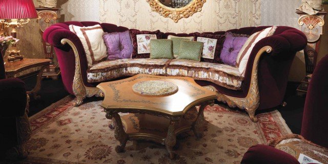 This image is of an ornately designed and decorated interior. It features a large, formal living room with several pieces of luxury furniture. There is a large red velvet sofa and two matching arm chairs set against a beige and golden wall, as well as a beautiful white and gold chandelier hanging from the ceiling. To the left of the sofa is an elaborately carved golden table and to the right is a lavish grey and gold bookshelf with several intricate figurines displayed on top.