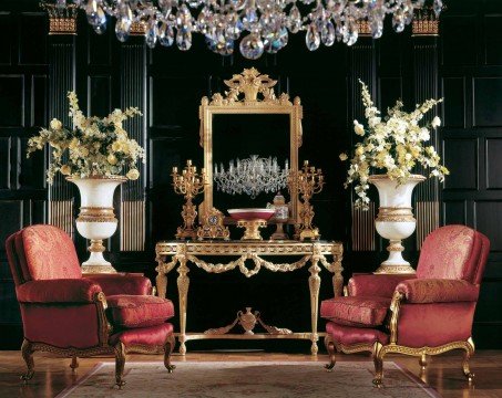 This picture shows a luxurious, modern staircase with a detailed gold railing and metallic accents. The steps are marble and the risers are painted a warm gold color. The walls around the staircase feature ornate white panels framed by narrow gold trim. A statement chandelier hangs from the ceiling, setting off the grandeur of the area.