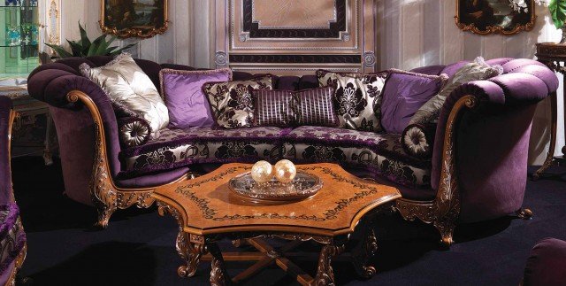 This picture shows a modern and luxurious living room. The room is filled with luxurious furniture and decorative pieces, including a white leather couch, two cream armchairs, a crystal chandelier, and two glass side tables. The walls are painted in a neutral beige color, and the floor is covered with a glossy, dark-colored tile. There is also a large beige area rug, and a set of double doors leading out to a patio.