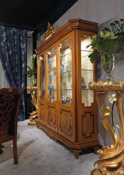 This picture is showing a luxurious marble staircase in an elegant home. The staircase has curved edges and ornate gold railings with intricate designs. There is a patterned tile floor with a beautiful rug in the middle of the stairs. At the top of the stairs is a stunning wall with an arch design, and there are two ornate chandeliers adding to the grandeur of the space.