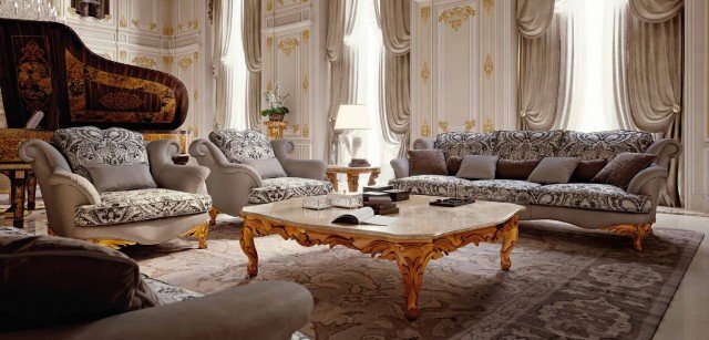 This picture shows an ornately decorated modern living room. The walls are white with gold accents, and cream and beige colors used for the furniture and carpet. A large white circular chandelier hangs from the ceiling and is surrounded by two wall sconces. The floor is covered with a dense white and beige patterned rug that contrasts the bright colors of the furniture. A large white sofa with intricate tufted details is paired with a tan leather armchair and a beige velvet ottoman, all framed by two ivory painted side tables with marble tops. On the walls