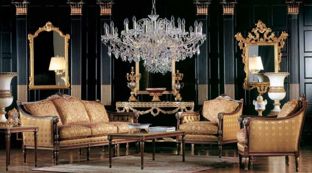 This picture shows a modern apartment living room featuring built-in wall units and furniture. The walls and floor are bright white and the furniture is upholstered in black. The furniture includes a sectional sofa, two arm chairs, and a glass top coffee table framed by an area rug. A large television mounted on the wall and two modern pendant lamps provide additional lighting. There are several black accent pieces in the room including a wall clock, a standing lamp, and a vase.