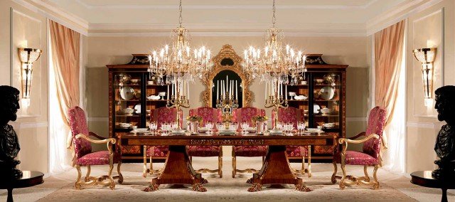 This picture shows a luxurious and opulent lobby in a modern hotel. The room has a high ceiling, with mirrored walls and white marble floors. The space is decorated with gold accents, including two large sofas and two round side tables, each with a tall golden lamp on top. At the back of the room is a white grand piano, surrounded by several ornate chairs.