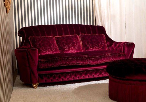 This picture shows a classic and elegant living room decor. The room features a white tufted sofa and two chairs, a glass-top coffee table, and a luxurious beige and gold rug. On the walls are framed artworks, black and brass lamps, and shelves filled with books, knick-knacks, and vases. The overall look is one of sophistication and style.