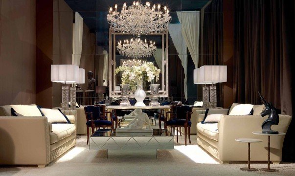 This picture shows an elegant modern entryway with an ornate ceiling and a curved staircase. The walls are decorated with mirrored wall panels, creating the illusion of more space. The floor is tiled in a light grey marble with a polished finish. A glass and metal balustrade lines the staircase and a grand chandelier is hung from the center of the ceiling. The whole effect is one of luxury and grandeur.