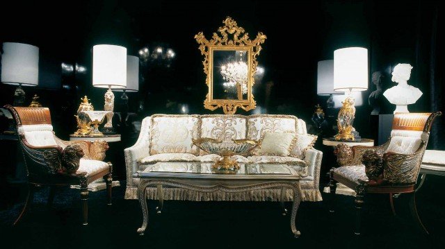 This picture shows a luxurious, modern bedroom with a light brown color palette. The central focus of the room is the large, upholstered bed, which features an eye-catching headboard and footboard. The walls are accented with gold trim, and on either side of the bed are matching nightstands with square mirrors above them. The other pieces of furniture in the room, such as the armchair, bench, and dresser, feature polished wood finishes and clean lines. Above the bed, there is a unique chandelier, adding to the sophisticated charm of the