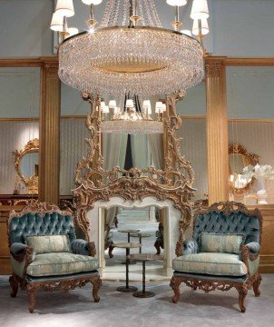 This picture shows a beautiful living room design with a unique modern aesthetic. The center of the room features an intricate brown and gold sofa, surrounded by two cream-colored armchairs and a matching ottoman. On either side of the seating area are tall cabinets, one black and the other white. The walls are decorated with wall art and a wooden shelf hangs over the sofa. There are also two end tables, a white rug, and a glass coffee table.