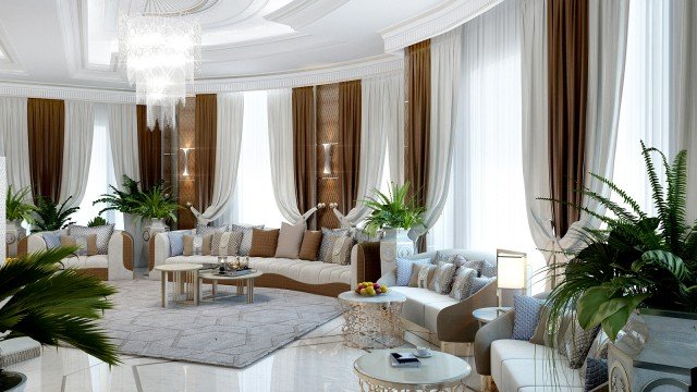 This picture shows a luxurious living room. The room has a large flat-screen TV mounted on the wall, two ornate armchairs upholstered in white, a cream-colored couch, and several beige and black accent pillows. An intricate area rug covers the dark hardwood floors, and a tall houseplant stands in the corner by the window. There is also a large wooden coffee table in the center of the room with several decorative items on top.