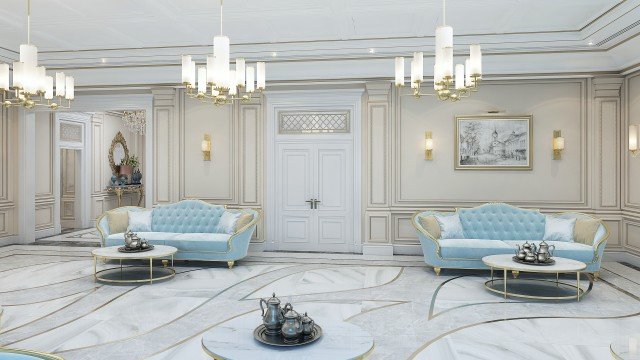 This picture shows an ornately-decorated interior room. The walls are painted in a soft gray color and feature elaborate gold detailing in the form of swirls and flowers. The floor is covered in a white and gray mosaic tile pattern. At the center of the room is a large white marble coffee table surrounded by several comfortable chairs, while two large chandeliers hang from the ceiling above.