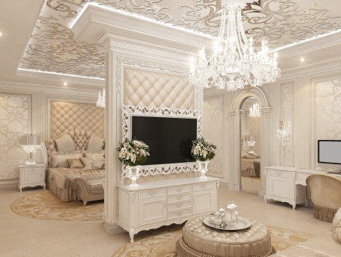 This picture shows a luxurious modern bedroom design. The room is decorated in shades of white and neutral colors with pops of gold accents. The walls are a light beige, and the floors and furniture are all white. The bed has a large, padded gold headboard with a quilted pattern. There are also two white side tables with intricate gold details, as well as two gold-framed mirrors on the wall. The ceiling is vaulted, and there is a large chandelier hanging from it. The room is finished with large curtains and a velvet bench at the end of