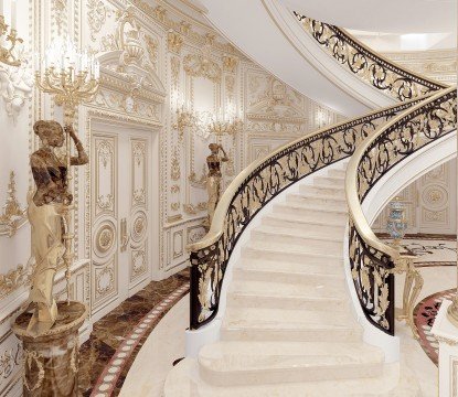 This picture shows an ornate gold and marble staircase in a luxurious home. The staircase is curved, with intricate gold details that contrast against the white marble steps and railings. The marble walls are decorated with gold accents and topped off with a luxurious crystal chandelier.