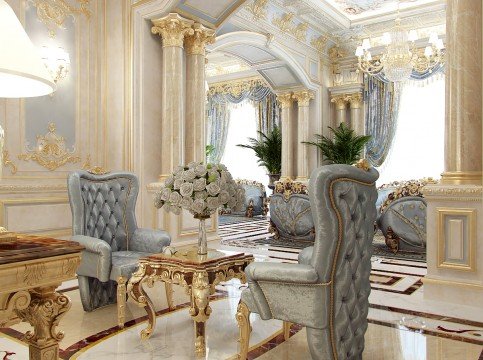 This picture shows an ornately decorated marble room. The room features a wrought iron railing on the curved stairway leading to a balcony overlooking the main room, white marble floors and walls with gold accents, and a white and gold ceiling decorated with intricate geometric patterns. There is a large carved marble fireplace with a floral-patterned marble mantle, and a luxurious crystal chandelier hangs from the ceiling. Furnishings in the room include two velvet armchairs, a gilded console table, and a large vase filled with white flowers.
