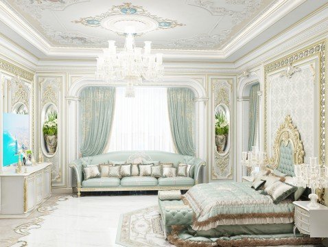 This picture shows a luxurious living room with high ceilings, marble flooring, and a plush white sofa with gold accents. The room is decorated with a large crystal chandelier and several wall lamps, along with a white and gold wall clock and a vase of white flowers. The walls are painted in an eggshell white color and are adorned with intricately patterned golden decorations.