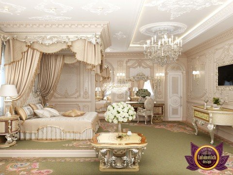 This picture shows an ornate, luxurious bedroom. It features a large, four-poster bed with a white frame and draped canopy, flanked by two bedside tables with golden lamps. The walls are a soft pink color with cream accents, and the floor is a rich-looking dark wood. In the corner is an armchair with decorative cushions in shades of blue and yellow, and on the walls are several pieces of art in gilded frames.