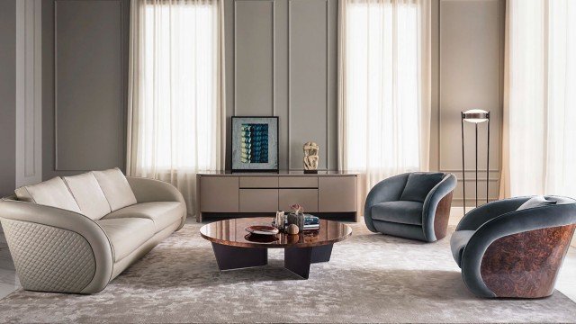 This picture shows a luxurious bedroom interior design. The walls are painted a light grey, and the floor is covered in a soft white shaggy rug. There is a large bed with a tall tufted headboard and a crisp white comforter. On either side of the bed are nightstands with decorative lamps. At the foot of the bed is a white velvet armchair and a black side table with a small, gold-plated vase. The ceiling has ornate crown molding, and there is a large window with sheer, cream-colored curtains.