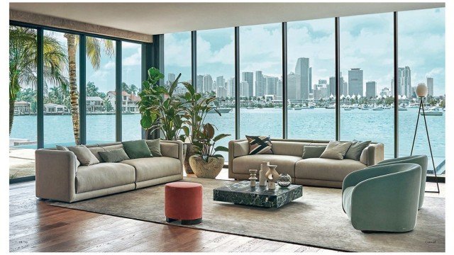 This picture shows an exquisite living room in a luxurious home. The walls are painted a light beige, and the ceiling is decorated with crown molding and a beautiful chandelier. In the center of the room is a large, comfortable looking cream-colored sofa, with four matching armchairs around it. There are two end tables next to the sofa, each with a lamp. On the left wall there is a fireplace, with built-in shelves and a flat-screen TV above it. On the right side of the room, there is a grand piano with an ornate chair