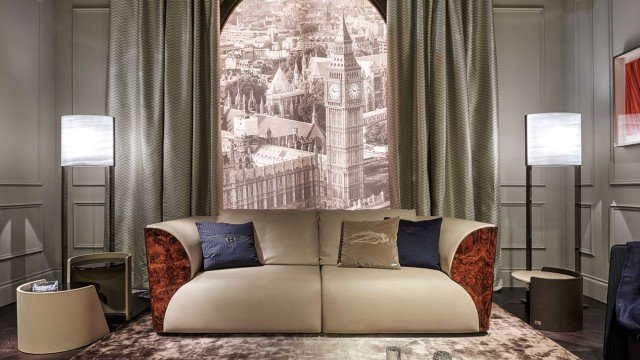 This picture shows a modern living room in an elegantly decorated apartment. The floor is covered in a light-colored wood and the walls are painted a deep golden hue. A large gray sectional couch runs across the far wall, adorned with sheer white drapes and colorful pillows. In front of the couch is a glass coffee table with a vase of flowers on top. A sleek flat screen television hangs on the wall above the couch. To the left of the couch is a low, mirrored side table with a lamp on top, and to the right is a tall bookshe