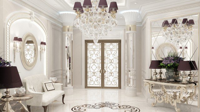 This picture shows a modern luxury foyer in a house. The foyer features a cream colored marble tiled floor, with an elegant marble staircase leading up to the second level of the home. There is an elaborate light fixture hanging from the ceiling, and a beautiful console table with a mirror on the wall. On the opposite wall is a cream colored settee and an accent chair in a plush velvet fabric, with a marble-topped end table next to it. The walls are accented with ornate crown molding and trim.