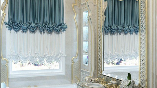 This picture is showing a luxurious bathroom with gold accents and marbled features. The room features a large bathtub with a chandelier above it, a white marble floor, a glass granite countertop, and a shower area with a built-in bench. The walls are lined with a beige tile and a gold accent border. The room is finished off with a glass vase filled with fresh flowers on the side.
