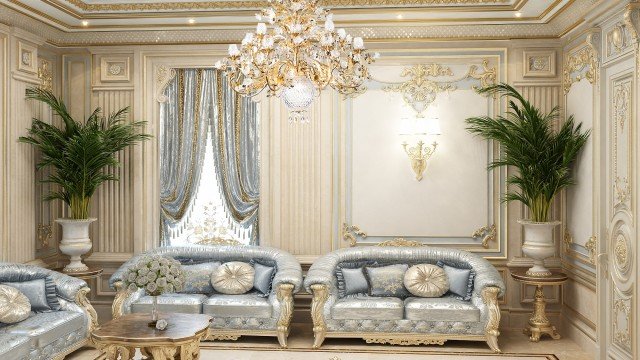 This picture shows a luxurious living room in warm, beige colors. It includes a large sofa with comfortable looking cushions and pillows, and several armchairs. A few large decorative plants are placed around the room, along with several small tables. A round carpet gives a cozy touch to the room. An ornate chandelier hangs from the ceiling, complementing the overall look.