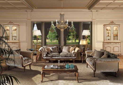 This picture shows a luxurious modern living room with white and grey walls, high ceilings, and a light wood floor. The room has a very large beige leather sectional couch with a wide rectangular coffee table and several coordinating armchairs in the same style. A white mantel with a television above it is mounted on the wall, as well as a few abstract art pieces. Two large sliding glass doors lead to a balcony which overlooks a park. On the left side of the room are two elegant marble-topped side tables with lamps and a decorative plant.