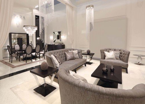 This picture shows an elegant interior design in the dining room of a luxury home. It has a large golden mirrored wall, a white marble table with eight modern black chairs, and two large glass chandeliers. The floor is made of dark wood and there is a luxurious rug in the center of the room.