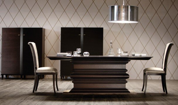 This picture is of an elegant dining room. The walls and ceiling feature an intricate geometric wallpaper, with a striped pattern in a bold color palette of white and gold. The room is illuminated by two ornate chandeliers. A grand wooden table is surrounded by six matching chairs with velvet upholstery. A large mirror hangs the wall above the table. On the far wall, there is a beautiful painting mounted onto the wall.