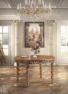 This picture depicts a luxurious open-plan living area. The room is decorated with modern white furniture, cream accents, and gold detailing. The seating area includes a leather couch and armchair with a golden coffee table, while the opposite side of the room features a dining space with a marble table and black leather chairs. The walls are painted a soft beige color and feature two large artistic paintings. The room is lit by a large crystal chandelier hanging from the white ceiling.