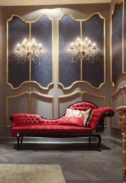 This picture shows a luxurious hotel suite with an ornate, modern design. The main feature is the sitting area which is composed of two plush couches in a satin gold color facing each other, centered around a trendy glass coffee table. The walls feature a dark gray and white chevron patterned wallpaper and are adorned with several pieces of contemporary art. In the foreground there is a regal armchair with a velvet paisley print fabric in green and cream colors. On the left of the image, there is a bed with a high headboard in a white, ivory and