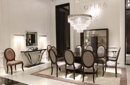 This picture shows a large, luxurious bedroom with elegant furniture and decor. The room is decorated in shades of grey and brown with hints of gold. The bed is large and ornate with a tufted headboard and matching bedding. A chandelier hangs from the ceiling, and an area rug covers the floor. Shelves line one wall and two armchairs are situated around a small table. Mirrors hang over the nightstands and a large window provides natural light.