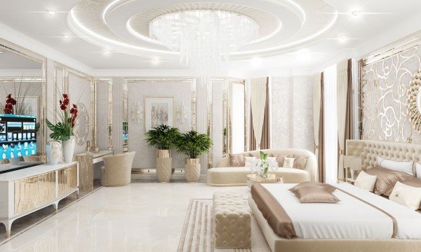This picture shows a luxurious living room with stylish and modern furniture. The room is elegantly decorated with white walls, elegant furniture, and a large round chandelier. The floors are dark-wood plank with an intricate pattern. The furniture is mostly upholstered in cream and light gray fabric with touches of gold throughout. The seating is composed of a three-seat sofa and two armchairs arranged around a low white coffee table. A grey and pink ottoman provides additional seating. A marble accent wall adds a luxurious touch to the room.