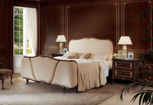 This picture shows a modern-style bedroom. The room is decorated in neutral shades of white, grey and brown. A large window with billowing curtains overlooks a view of a lake and lush greenery outside. A bed with a simple, neutral-colored headboard and bedding is centered in the room. A matching nightstand sits next to it, and a geometric patterned rug covers the floor. A long, upholstered bench is at the foot of the bed, while an ornate mirror is hung on the wall above it.