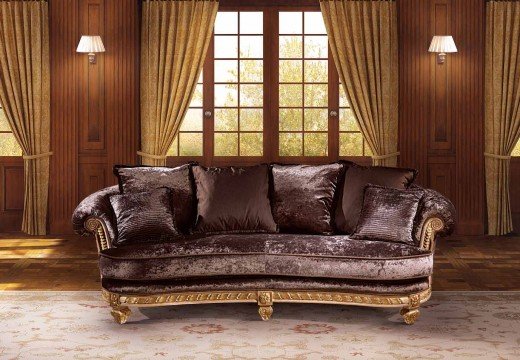 This picture shows a modern living room with an elegant, minimalist design. The walls are painted a neutral beige color and the floor is covered in a light beige carpet. There is a grey sectional sofa with a couple of white accent pillows and a rectangular coffee table with a glass top. On one side of the room is a low bookshelf with several decorative books and a few framed photographs. In the corner is an armchair with a round, metal side table next to it. A bright landscape painting is hung on the wall above the couch, adding a splash of color