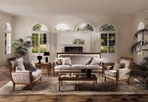 This picture shows a luxurious living room with a large sectional couch upholstered in a light tan velvet. The walls are a dramatic deep navy blue color, accented by white and gold detailing on the ceiling and around the room. A black marble fireplace adorns one wall and the flooring is a striking white and black checkered pattern. Several glass doors lead out to a terrace area. An elegant chandelier hangs from the ceiling, accentuating the grandeur of the space.