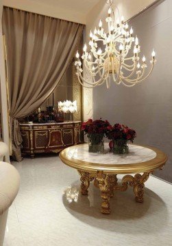 This picture shows a modern and luxurious living room. The walls feature off-white paneling with gold accents, while the floor is made of light wooden planks. There are two large white sofas with ornate patterned pillows, and a sleek glass coffee table between them. In the corner of the room is a white grand piano with a black bench, and behind it is a bookshelf filled with books and framed artwork. The furniture is accented with several pieces of modern decor, including a long gold mirror and a white vase with a bouquet of fresh flowers