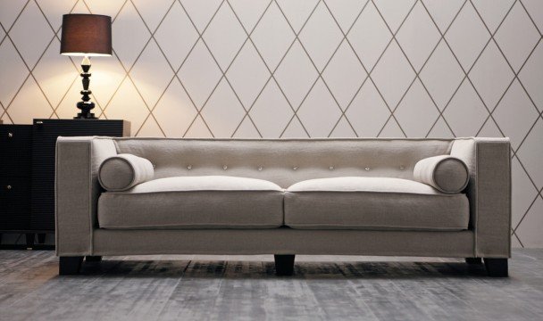 This picture shows a living room with two white sofas and two armchairs arranged around a coffee table. The furniture is set on a beige carpet in front of a large blue wall with a row of built-in open shelving. The shelves have decorative accessories such as vases, plants, books, and framed artwork. A large chandelier hangs from the ceiling, and there are several windows that allow natural light to enter the room.